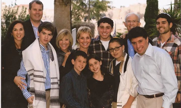 Twelve people of several generations crowded together for a group photograph outdoors, stone buildings and trees behind them, two of them wearing prayer shawls.