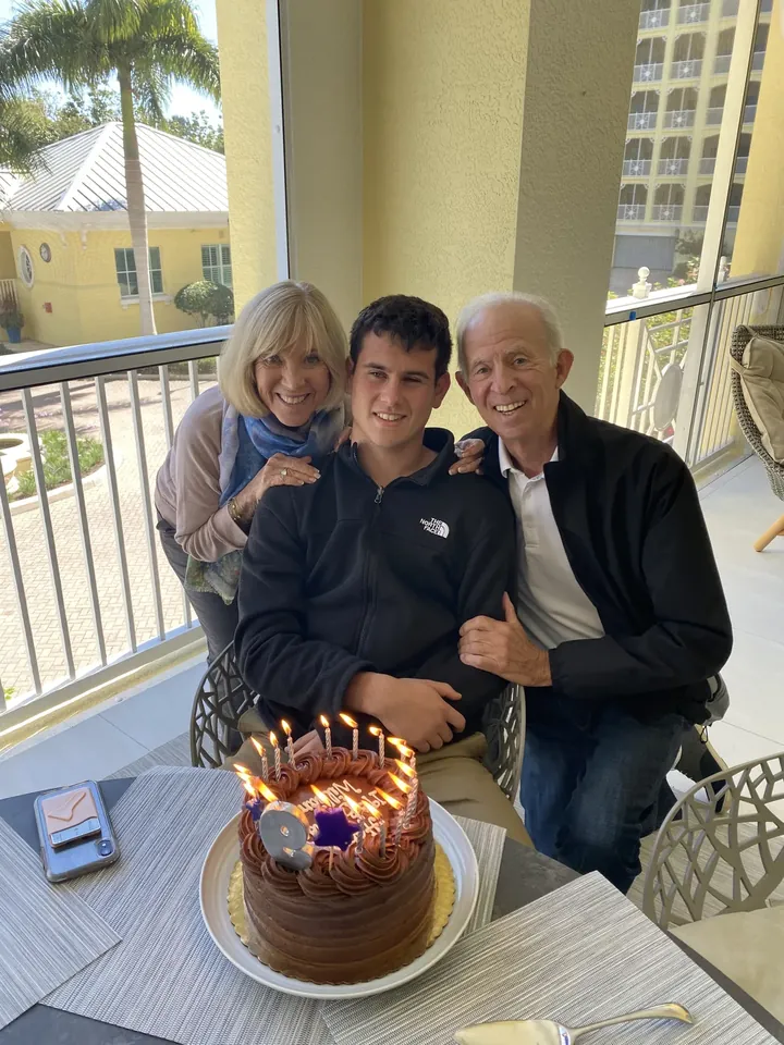 A young man seated between an older woman and an older man on a balcony, a lit chocolate cake on the table in front of them.