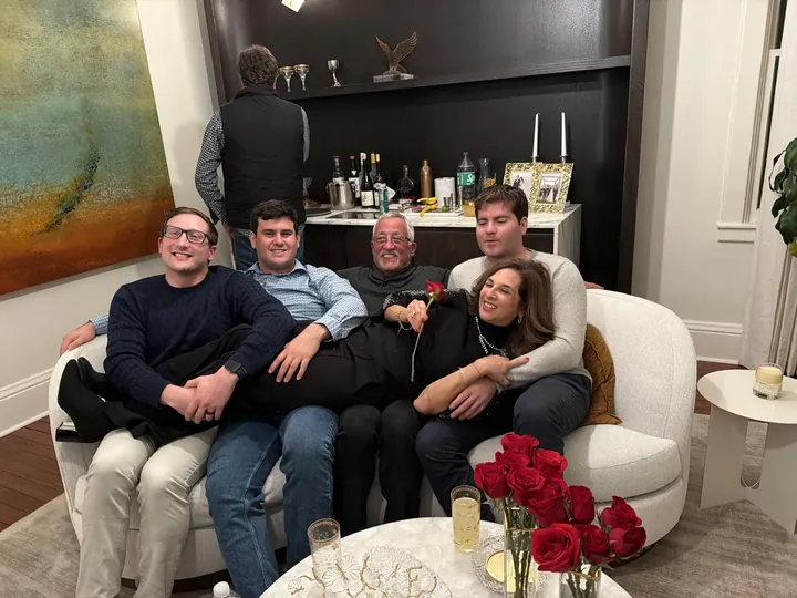 A group of people piled together on a white sofa, laughing, a vase of red roses on the table in front of them.
