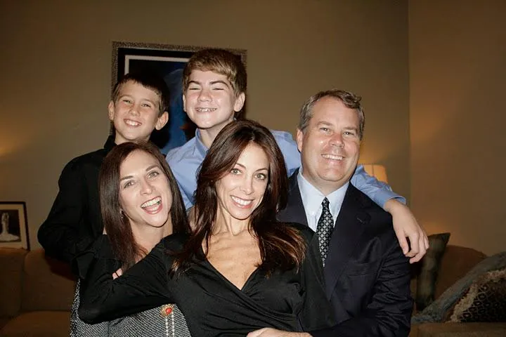 Two boys, two women and a man crowded together on a sofa in a lamp-lit living room, all laughing at the camera.