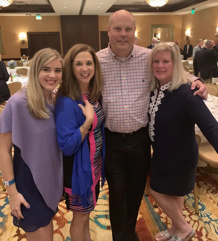Four people standing arm in arm in a hotel ballroom, a young woman, two women and a man in a checked shirt, laid tables behind them.