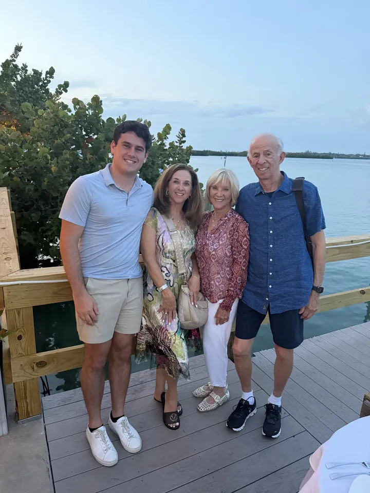 A young man, two women, and an older man standing together on a wooden dock at dusk, calm water behind them.