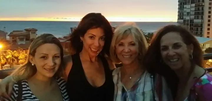 Four women standing shoulder to shoulder on a balcony at sunset, the sea and a low skyline behind them.