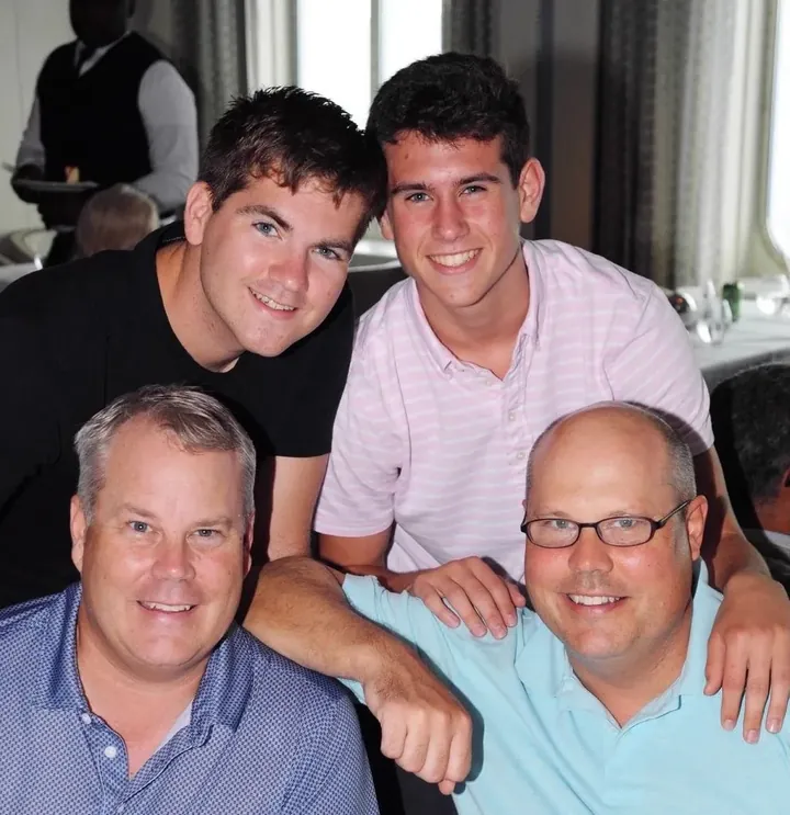 Four people crowded together at a restaurant table, two younger men leaning in behind two older men, all smiling.