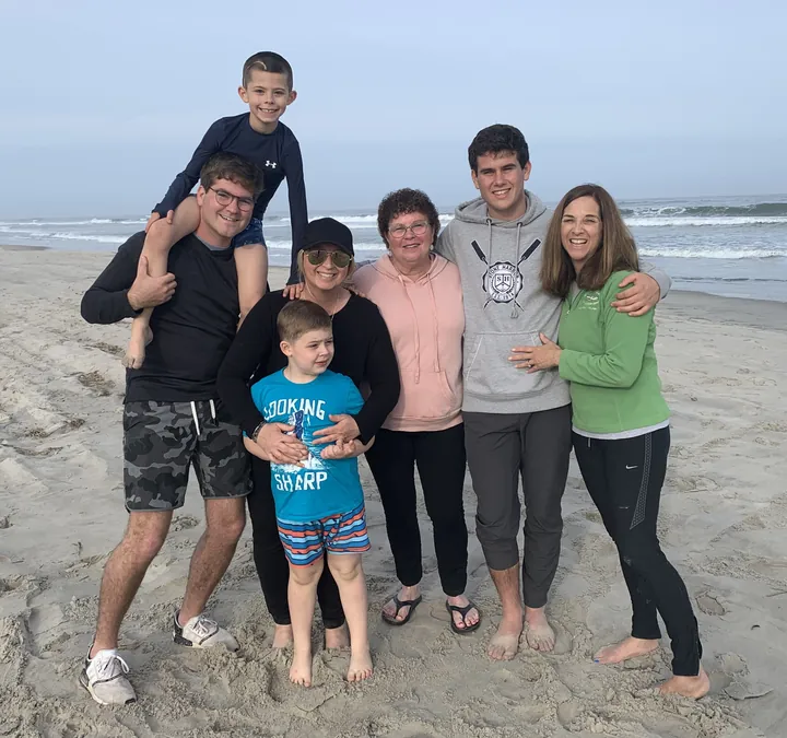 Seven people standing together on a wide grey beach, a boy up on a man's shoulders and a small boy in a blue t-shirt in front, the surf behind them.