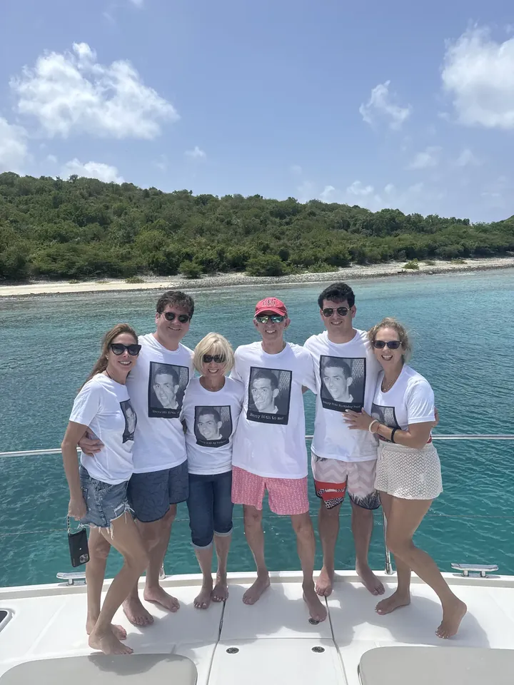 Six people standing barefoot on the bow of a boat, all wearing matching white t-shirts printed with a man's face, a green island shore behind them.