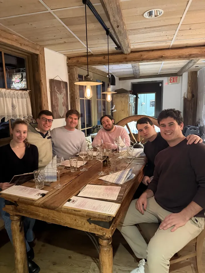 Six people gathered around a long wooden table set with wine glasses and menus, smiling toward the camera.