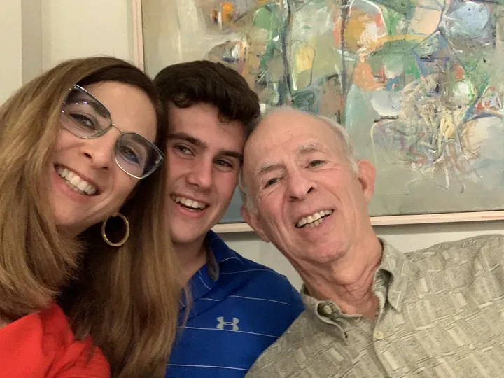 A woman in glasses, a young man, and an older man pressed cheek to cheek for a photo, all laughing, an abstract painting behind them.
