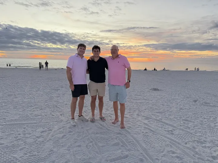 Three men standing side by side on wet sand at the water's edge, a colourful sunset behind them.