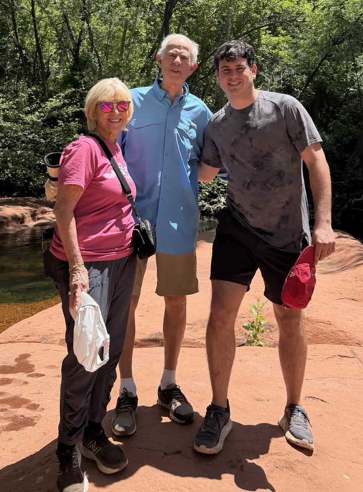 An older woman in a pink t-shirt, an older man in a blue shirt and a young man standing together on red rock beside a creek, green woods behind them.