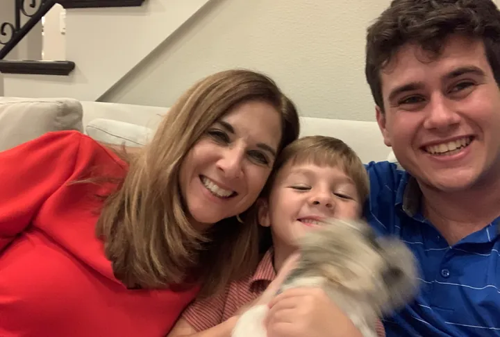 A woman, a small boy and a young man pressed together on a pale sofa, all laughing, a small dog held up in front of them.