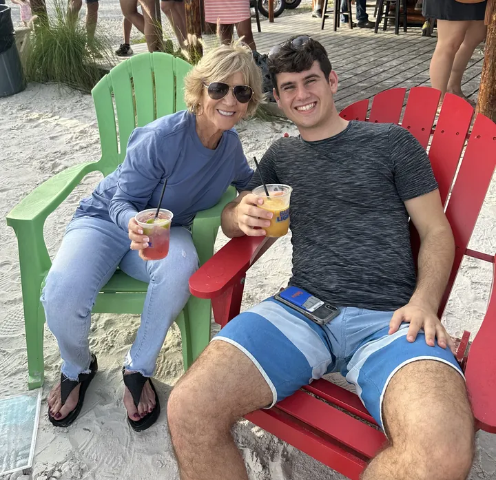 An older woman in sunglasses and a young man sitting back in green and red beach chairs on the sand, each raising a drink.