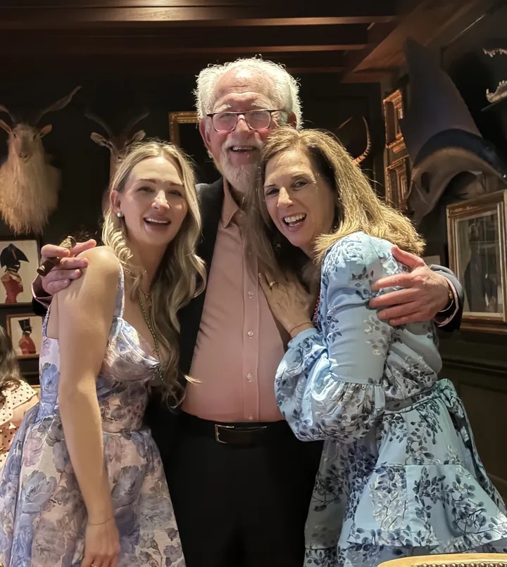 An older man with a white beard and glasses standing between two women, all three laughing, framed pictures and mounted animal heads on the wall behind.