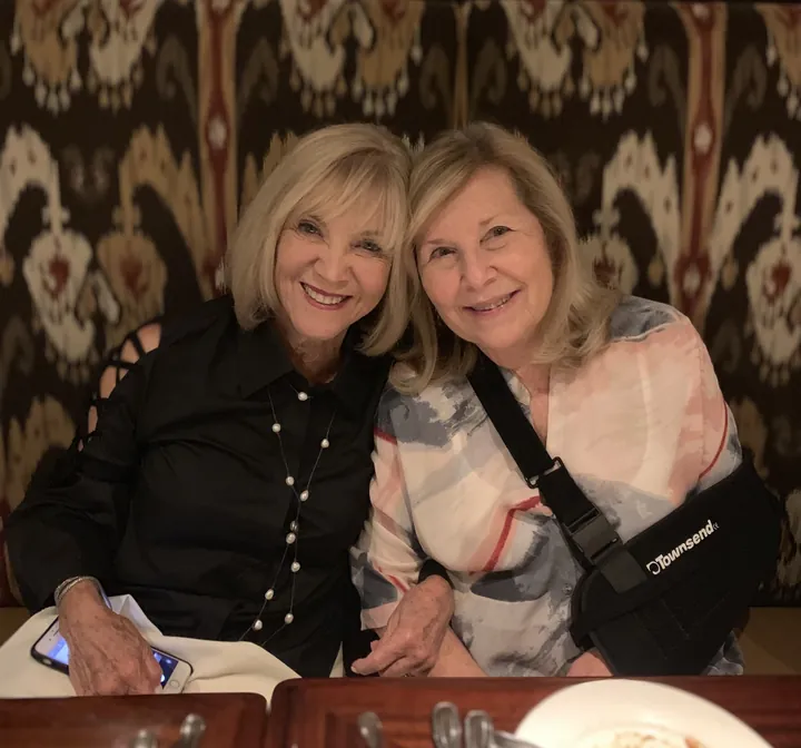 Two older women with blonde hair leaning their heads together in a restaurant booth, one wearing a sling on her arm.