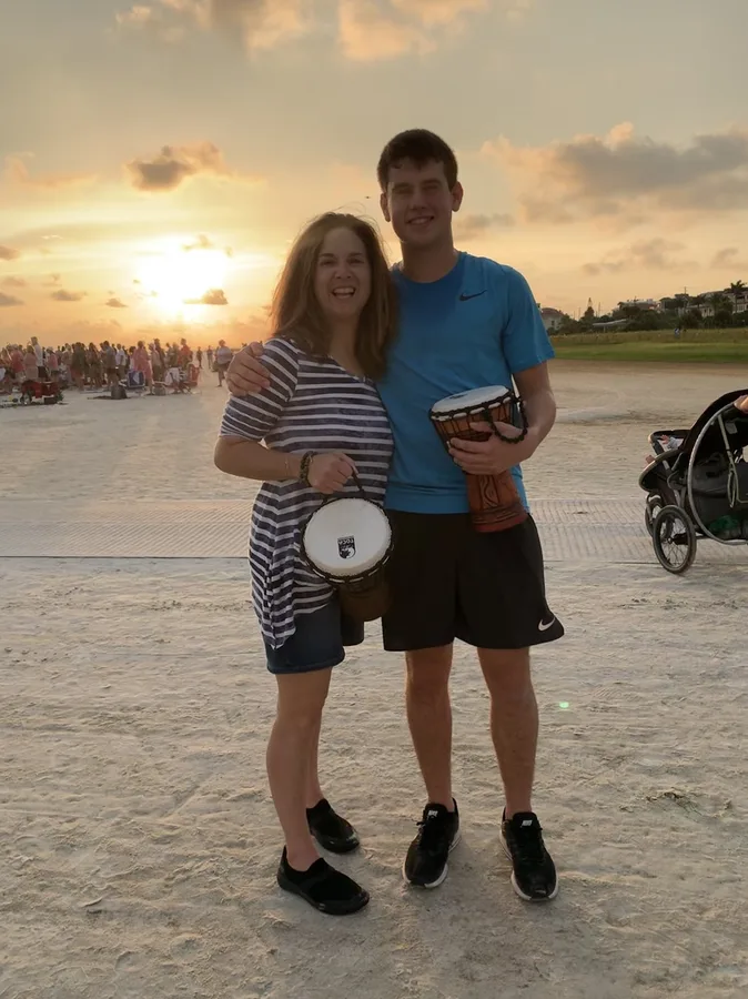 A woman and a young man standing together on the sand, each holding a hand drum, the sun low behind them.