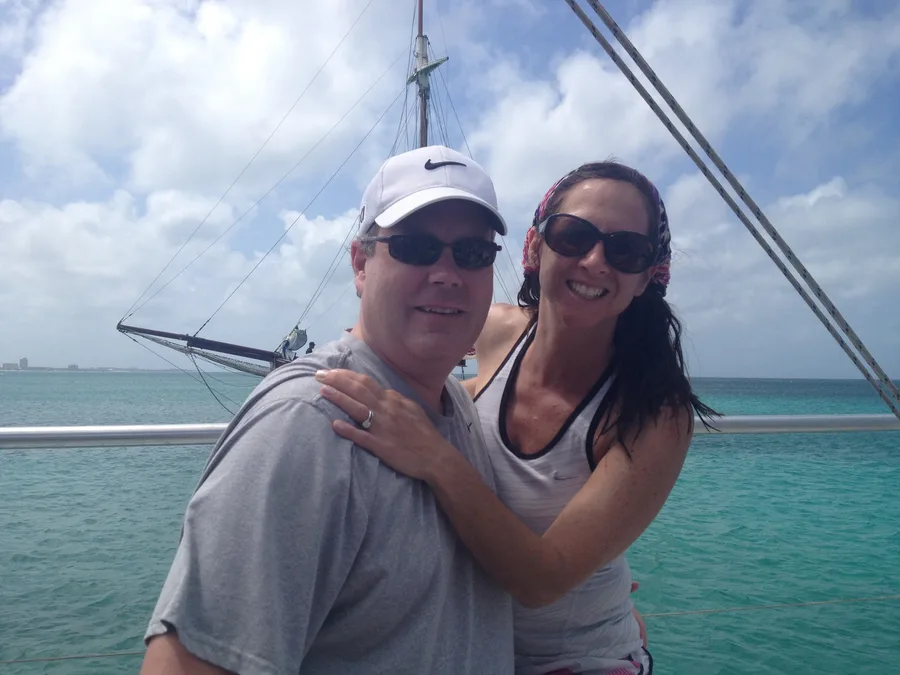 A man in a cap and a woman, both in sunglasses, leaning together on the deck of a sailboat, turquoise water behind them.